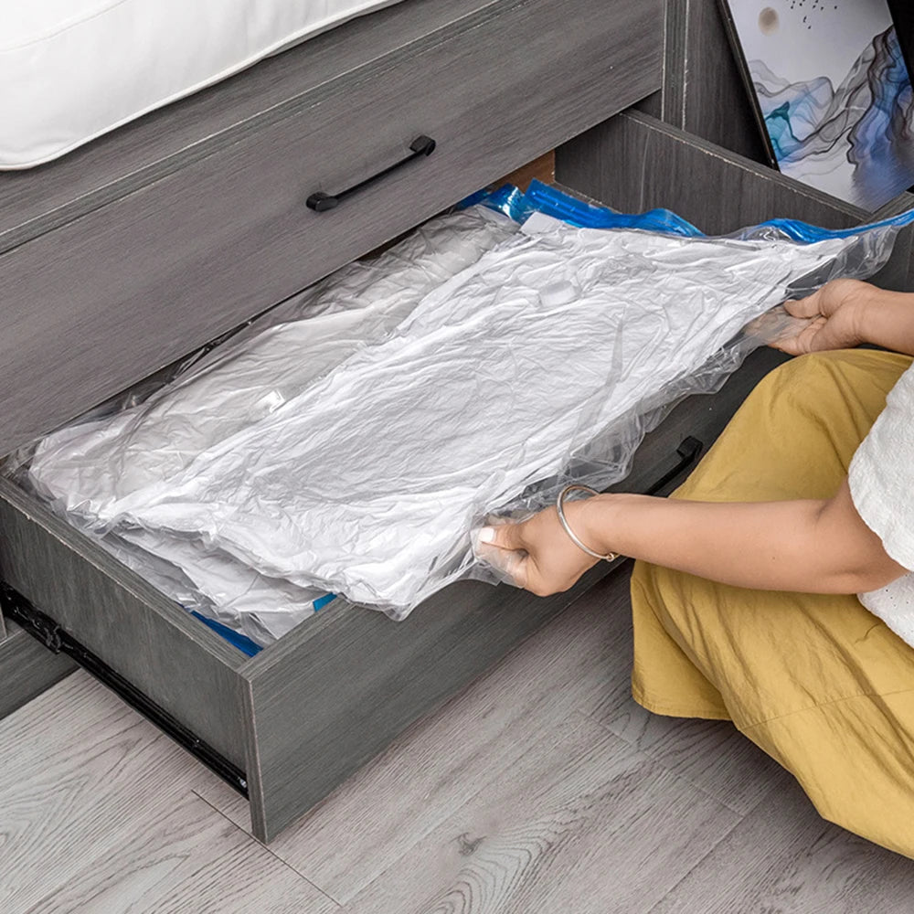 Person storing vacuum-sealed white bedding in gray under-bed storage drawer on wood floor